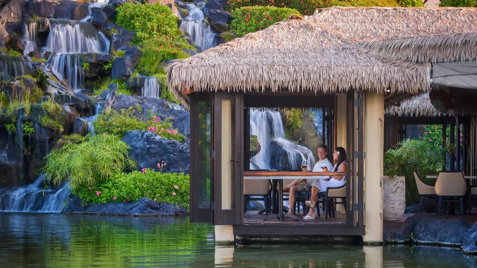 A couple dining under a palapa in Hawaii in front of waterfalls.