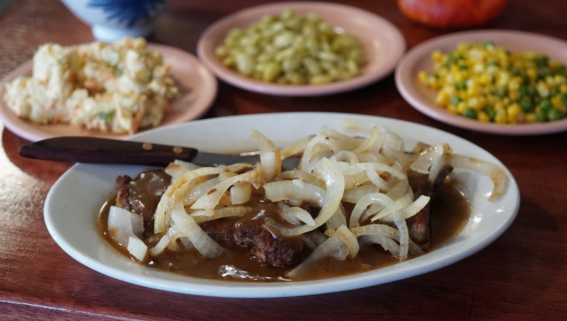 Table full of food, pork chops with onions, peas and corn, coleslaw.